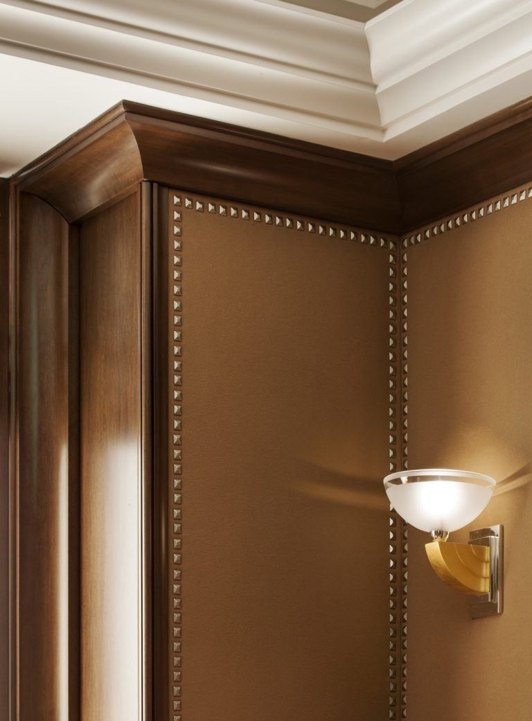A close-up of a corner in an elegant room with brown, leather-textured walls accented by metallic studs, dark wood paneling, detailed crown molding, and a modern wall sconce with a frosted glass shade and gold arm.