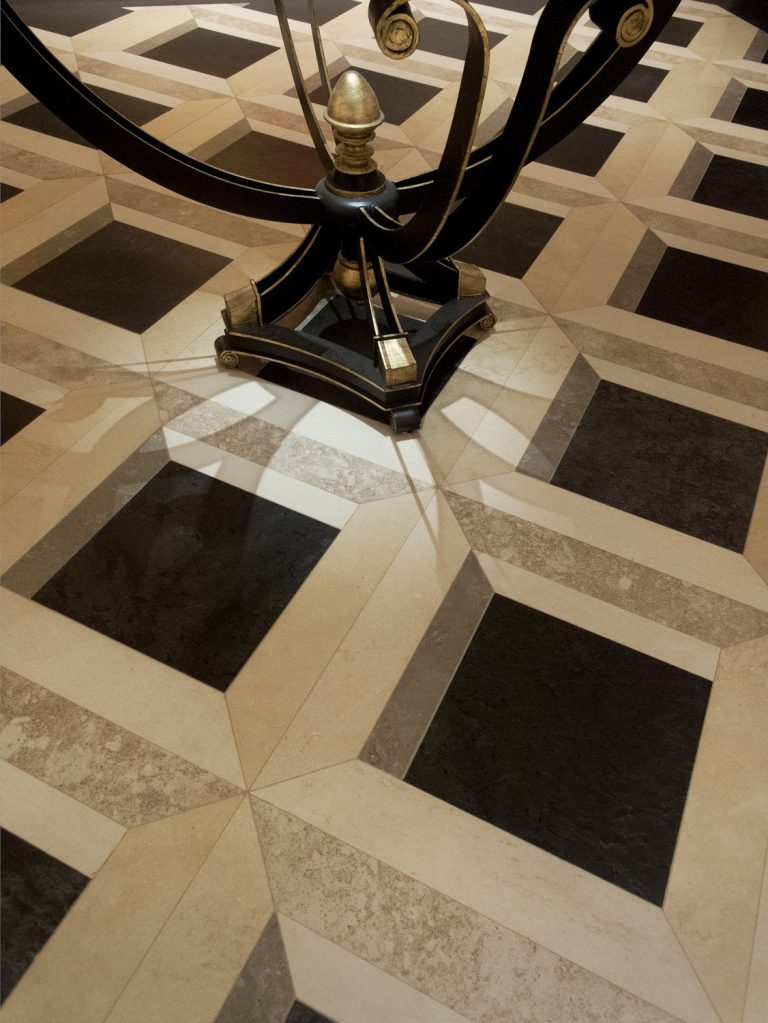 A geometric tile floor with a 3D cube pattern in black, beige, and tan tones. The ornate black and gold base of a metal table stands on the tiles, casting soft shadows and enhancing the illusion of depth.