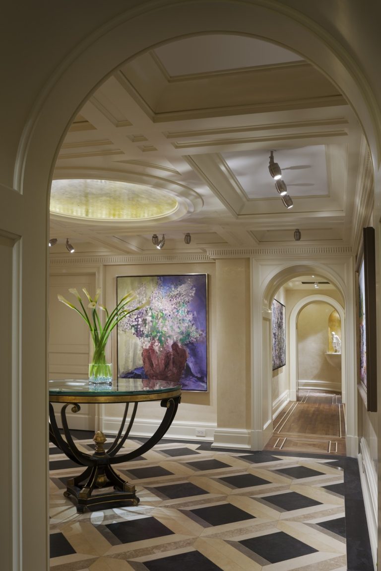 Elegant hallway with an arched entry, coffered ceiling, track lighting, and checkered marble floor. A round glass table with a vase of long-stemmed flowers is centered; vibrant floral paintings hang on the cream-colored walls along the corridor.