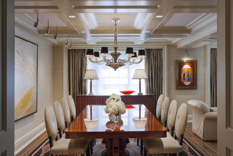 A formal dining room with ten upholstered chairs around a polished wood table, topped with white flower arrangements and bowls. A modern chandelier hangs above. Large art pieces adorn the walls, and windows with drapes let in natural light.