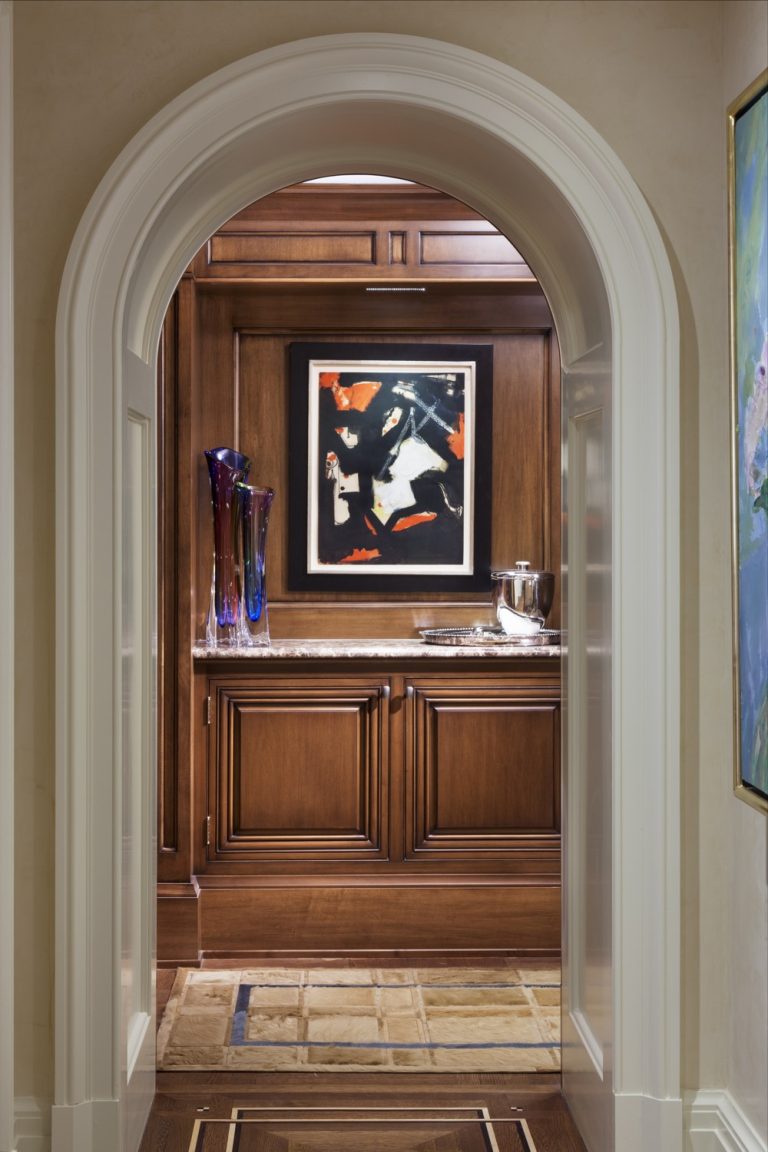 A view through a white-arched doorway reveals a small room with wooden cabinets, abstract art on the wall, tall colorful glass vases, a metal ice bucket, and a tiled floor. The space appears elegant and warmly lit.