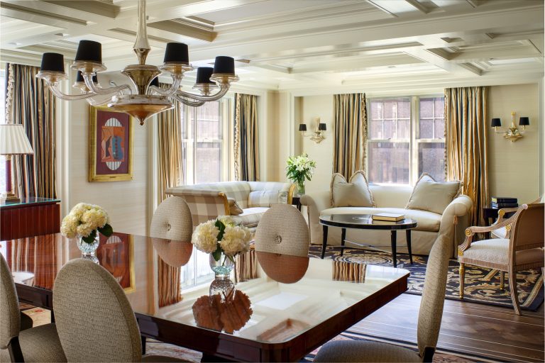 Elegant living-dining room with beige walls, coffered ceiling, and wood floors. Cream sofas, gold and black drapes, a chandelier, fresh flowers, and artwork decorate the space. Large windows let in natural light. Dining table with upholstered chairs is in the foreground.