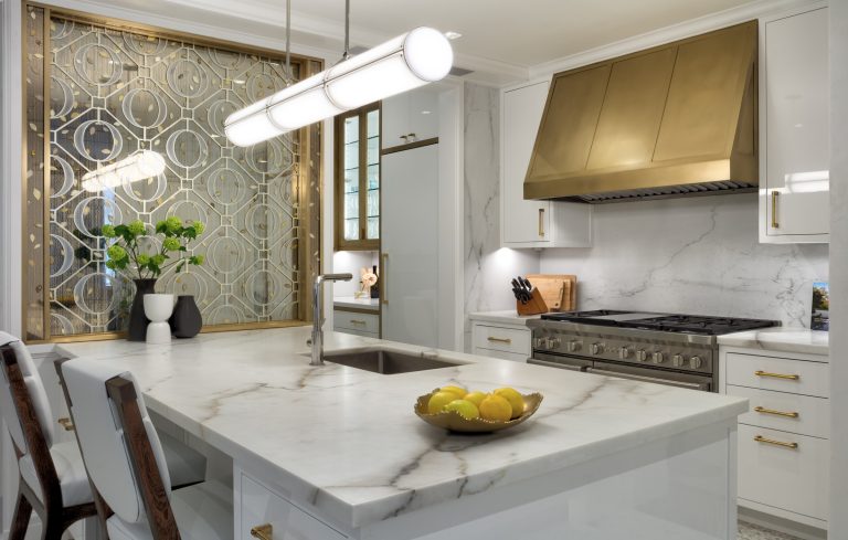 Modern Park Ave Apartment kitchen with white marble countertops, gold accents, and a brass range hood. A mirrored wall, white cabinets, and fresh flowers near elegant pendant lighting create classic New York sophistication.