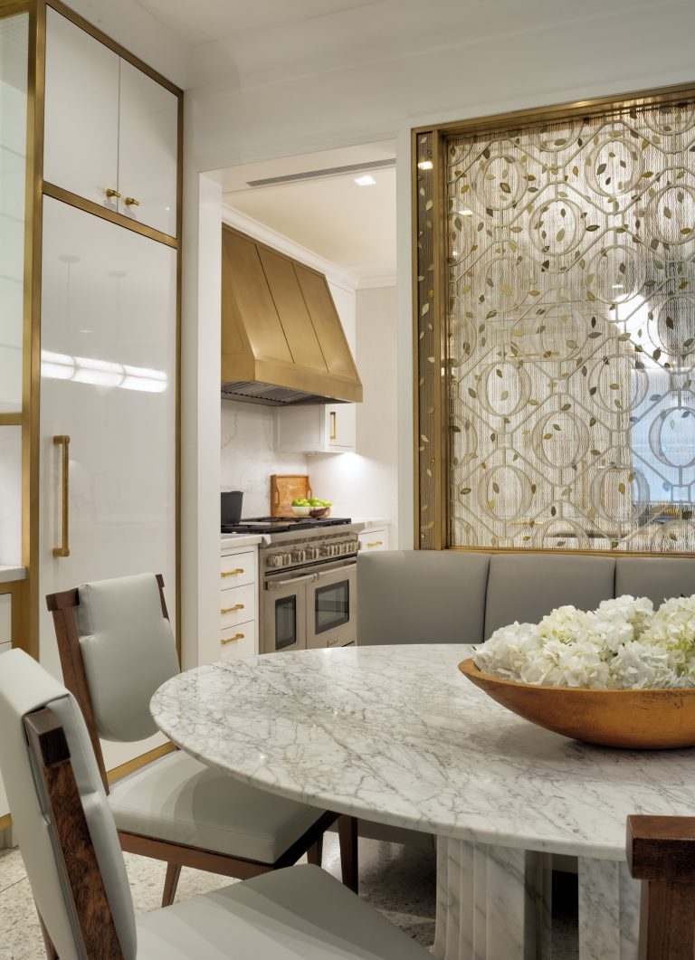 A modern Park Ave Apartment kitchen in New York features white and gold cabinets, a gold range hood, and a marble-topped round dining table. A golden decorative screen and a bowl of white flowers add elegance to the contemporary space.