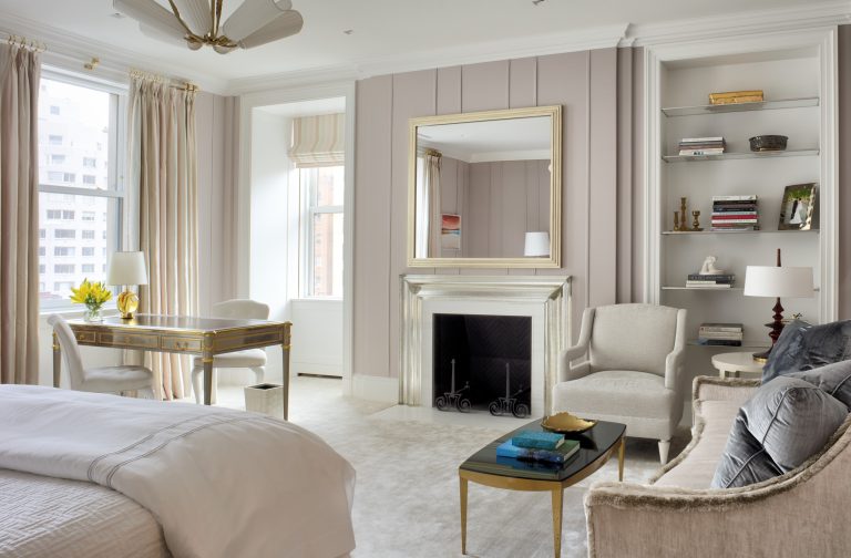 Elegant, bright Park Ave Apartment bedroom with neutral tones, large windows, a gold-trimmed desk with flowers, fireplace and mirror, built-in bookshelves, upholstered chairs, coffee table, and a neatly made bed with crisp white bedding.