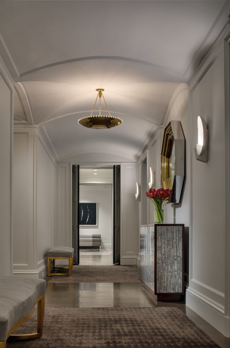 A modern Park Ave Apartment hallway with light gray walls, a beige carpet runner, and a cushioned bench on the left. A console table holds red tulips under a gold-trimmed mirror. Wall sconces and a circular chandelier softly light the space.