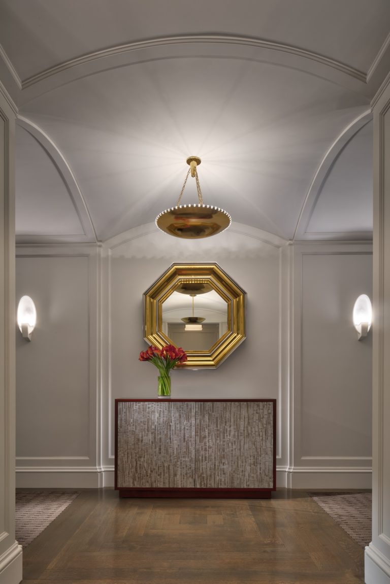 An elegant Park Ave Apartment hallway with an arched ceiling, a gold octagonal mirror above a marble console table holding red flowers. A round gold chandelier hangs above, while sconces softly light beige paneled walls and the wooden floor.
