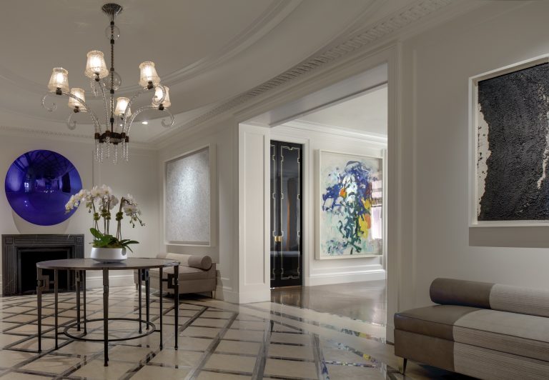 Elegant Park Ave Apartment foyer with a chandelier, round table holding white orchids, and geometric-patterned floor. Modern abstract art adorns cream walls; a large reflective purple circle crowns the fireplace. Hallway displays more New York artworks.