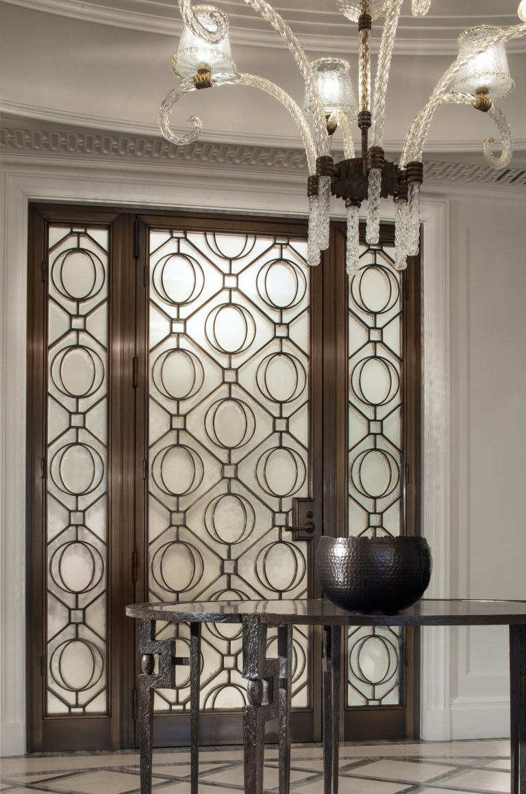 Elegant entryway in a Park Ave Apartment with double doors featuring intricate geometric glass and metal patterns. A glass chandelier with curved arms hangs above a modern metallic table, elevating the sophisticated New York space.