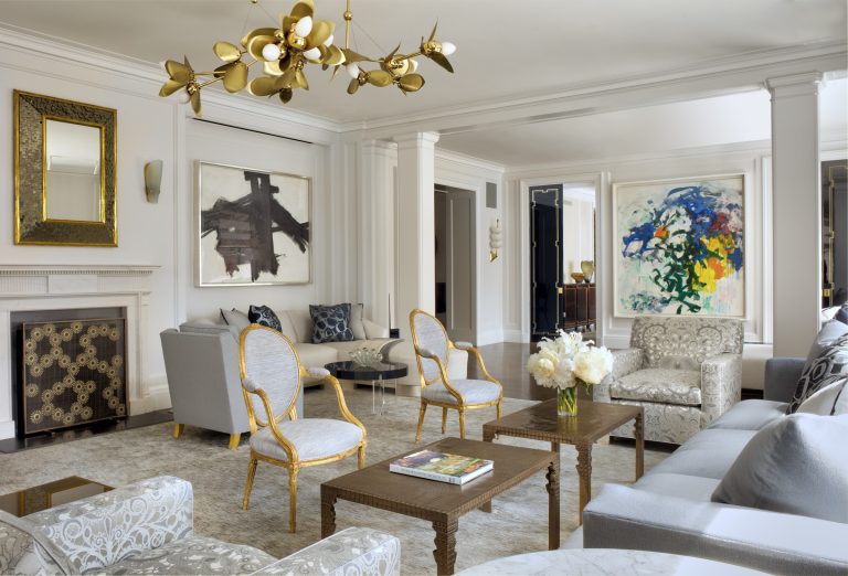 Elegant Park Ave Apartment living room with neutral tones, gold accents, classic armchairs, gray sofas, marble fireplace, abstract wall art, and a modern gold chandelier. Large windows fill the New York space with natural light for a bright, airy feel.