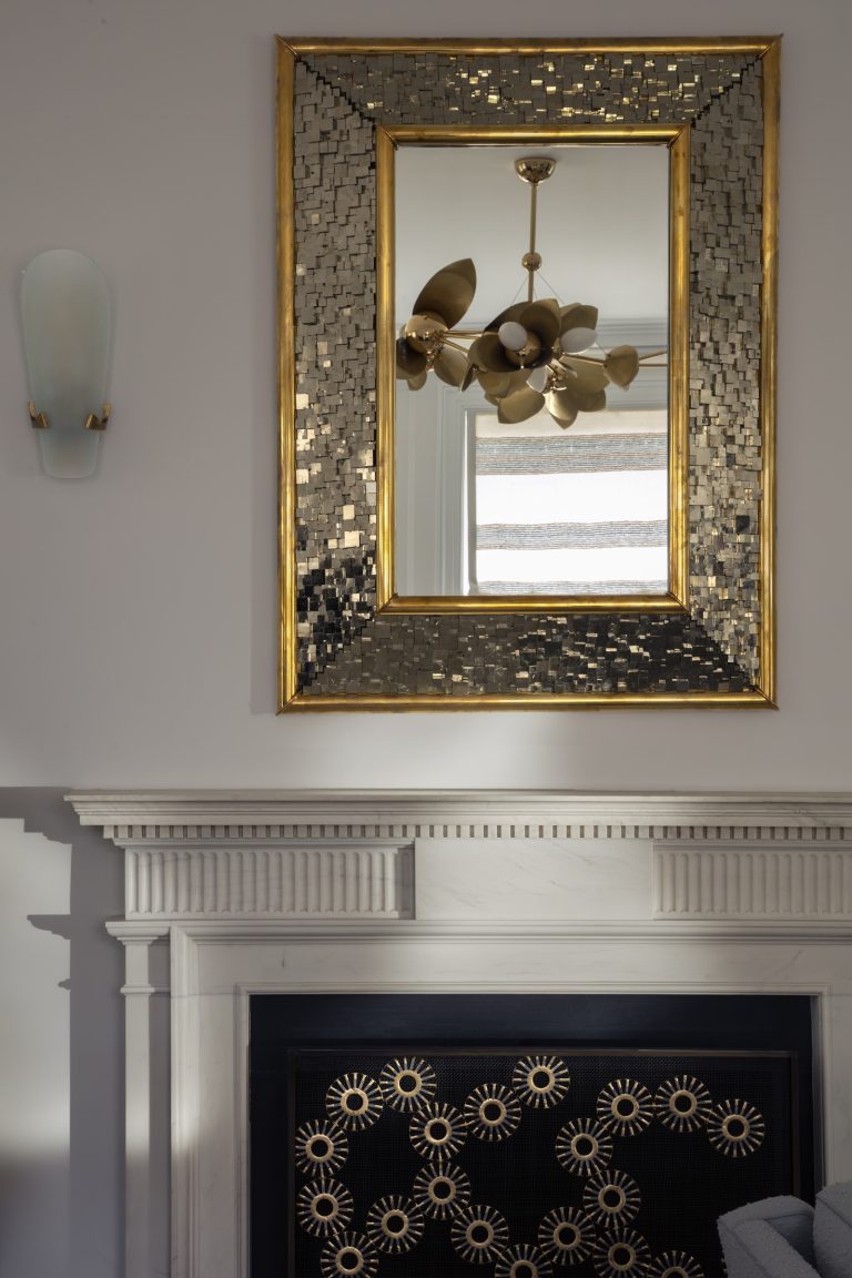 A gold-framed, mosaic-style mirror hangs above a white mantelpiece in this Park Ave Apartment. The mirror reflects a gold modern chandelier and a window with a striped blind, echoing classic New York elegance.
