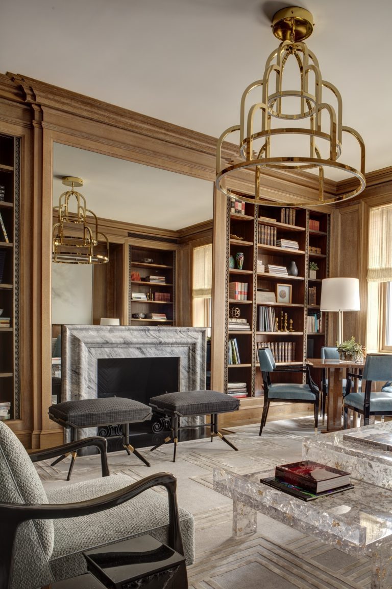 Elegant Park Ave Apartment library with wood-paneled walls, large mirror, marble fireplace, two dark chairs, blue armchairs, bookshelves filled with books and decor, a tall lamp, marble coffee tables, and gold chandeliers. Sunlight streams through tall windows.