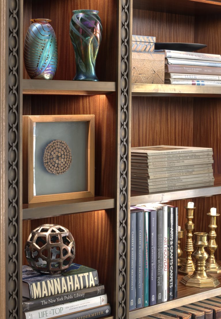 A close-up of a wooden bookshelf in a Park Ave Apartment displays colorful vases, stacked books, a framed geometric art piece, a bronze geometric sculpture, and several gold candlesticks. The shelves are neatly arranged and warmly lit.