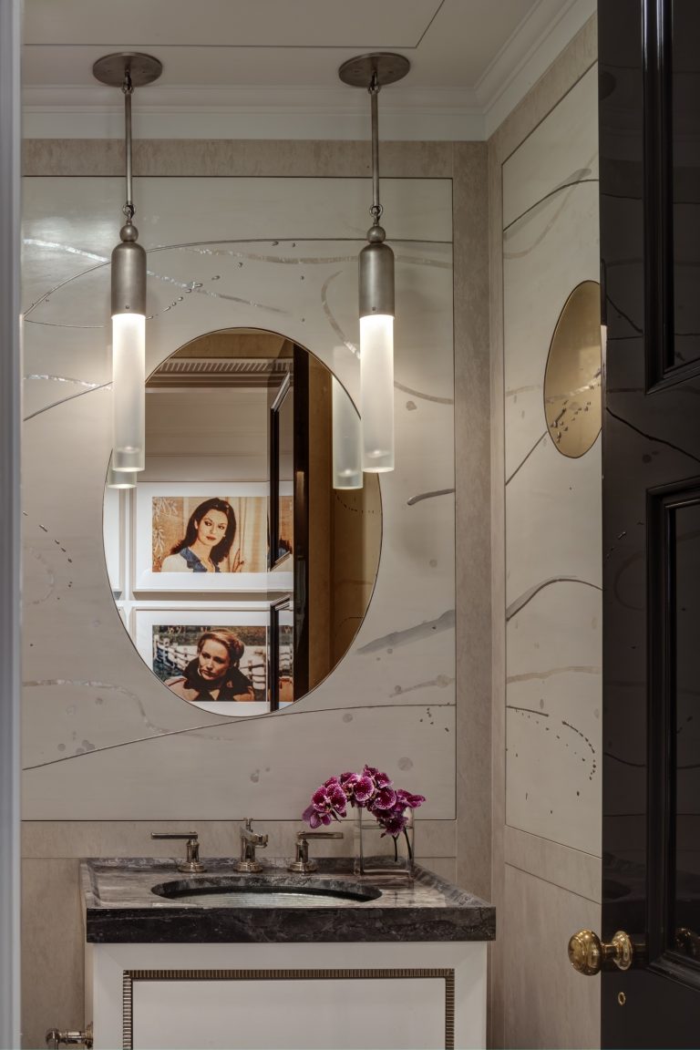 A bathroom vanity in a Park Ave Apartment features a black marble countertop, silver faucet, and pink orchids. An oval mirror reflects framed portraits while pendant lights hang from the ceiling, all set against light paneling with metallic accents.