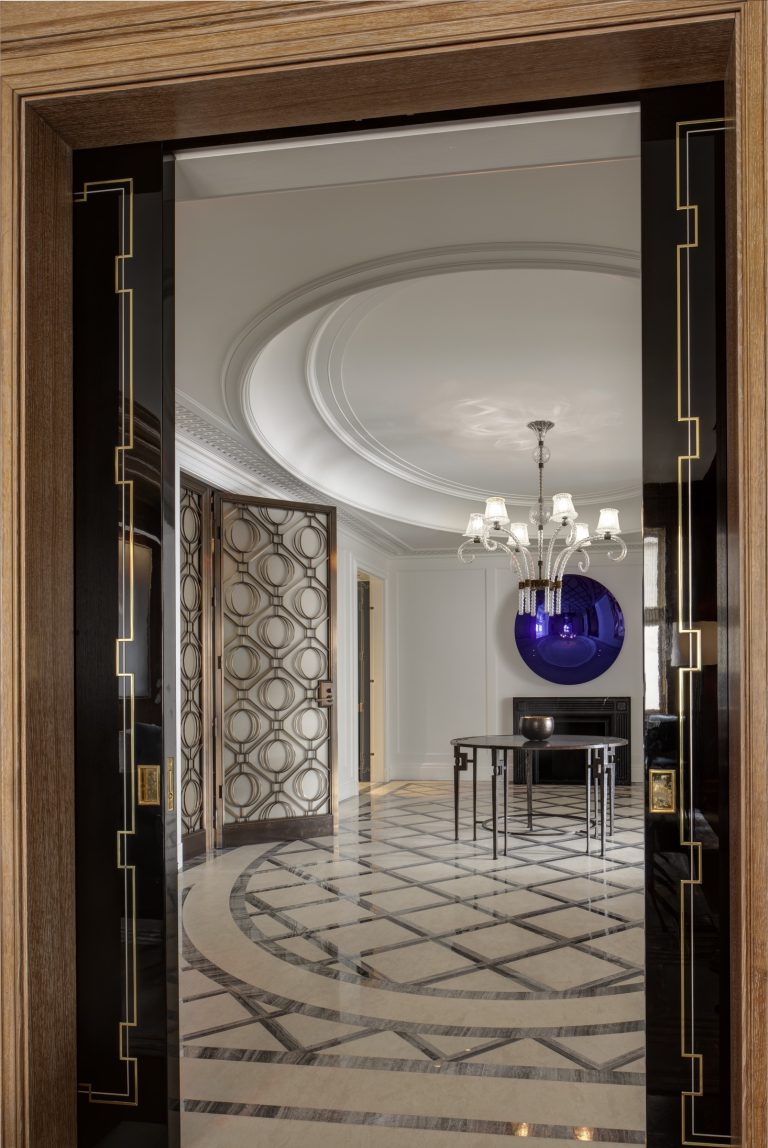 View through a doorway into an elegant Park Ave Apartment in New York, featuring geometric marble flooring, a round black dining table, modern chairs, a dramatic purple wall sculpture, ornate ceiling molding, and a multi-light chandelier.