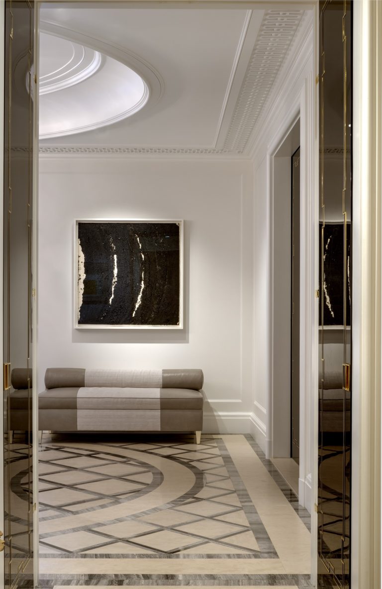 Elegant hallway in a Park Ave Apartment with patterned marble floor, cream walls, and ornate molding. A modern abstract painting hangs above a sleek, striped bench. Soft natural light enters from an oval ceiling skylight in New York.