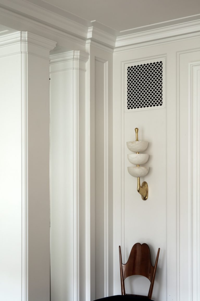 A minimalist, elegant corner of a Park Ave Apartment in New York features white paneled walls, a sculptural wall sconce with three bowl-like shades and brass accents above a vintage wooden chair. A black HVAC vent sits above the light fixture.