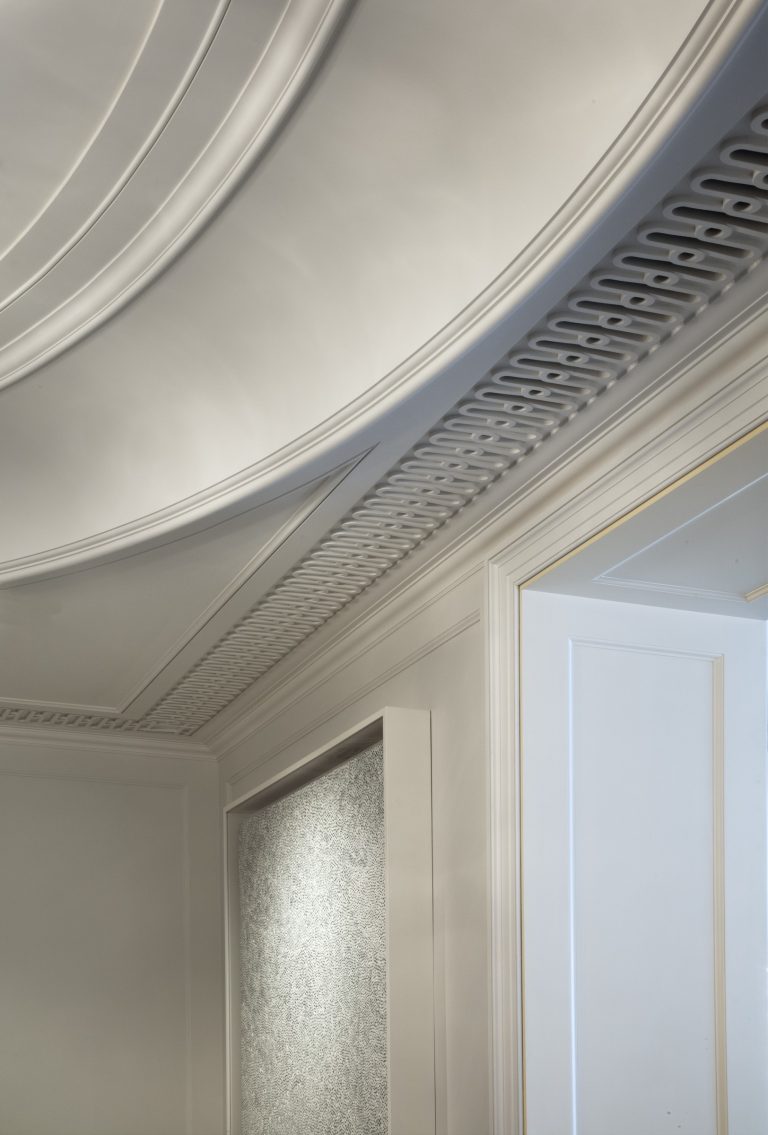 Curved white ceiling with detailed molding and vent grilles, above a textured gray wall panel and a doorway with smooth, minimalist trim. Soft, natural light highlights the sophisticated design of this Park Ave Apartment in New York.
