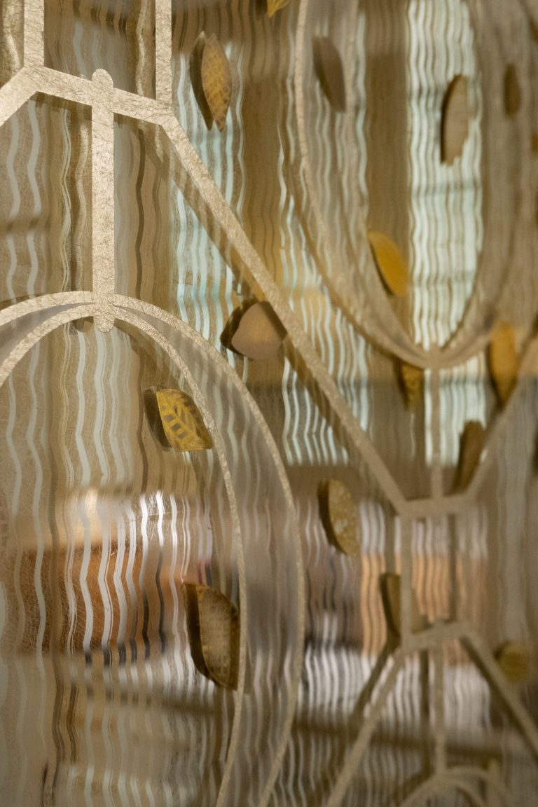A close-up of decorative frosted glass with a wavy, textured surface in a Park Ave Apartment. The glass features a gold geometric pattern with leaves, and soft, blurred colors can be seen through the glass in the background.