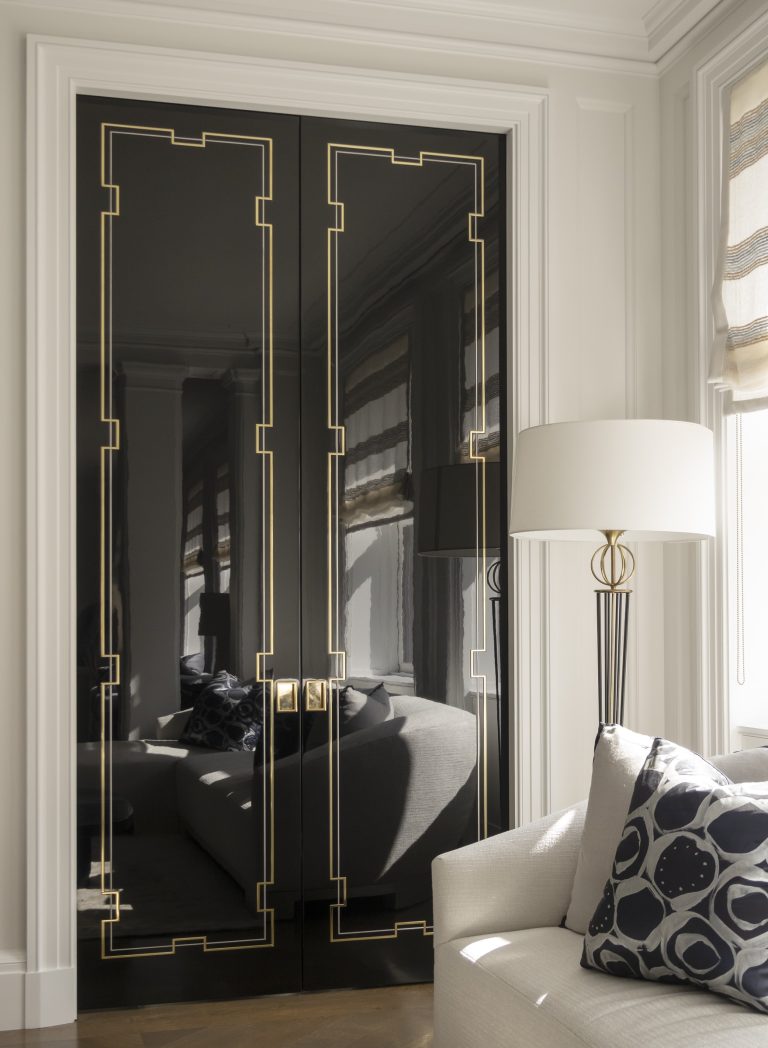 A modern Park Ave Apartment living room in New York, with glossy black double doors featuring gold geometric trim, reflecting a window and gray sofa. A white floor lamp with gold accents and a white armchair add elegance to the space.