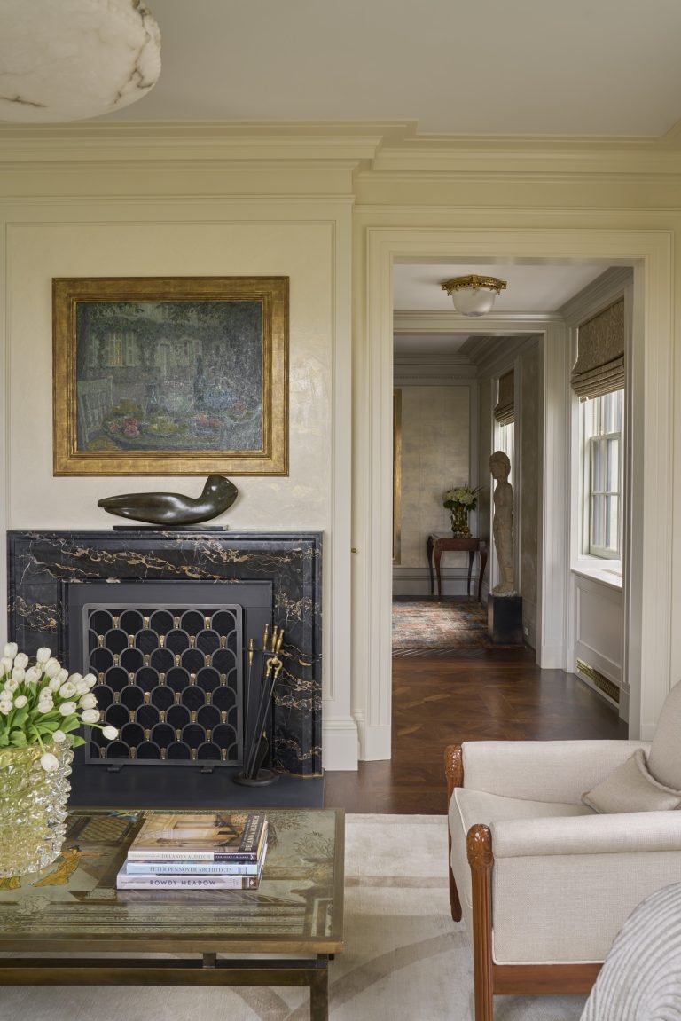 Elegant living room in a New York Fifth Avenue renovation with a black marble fireplace, gold decor, and modern sculpture. Framed painting above, glass coffee table with books and white tulips. Hallway with wood floors and a sculpture visible beyond.
