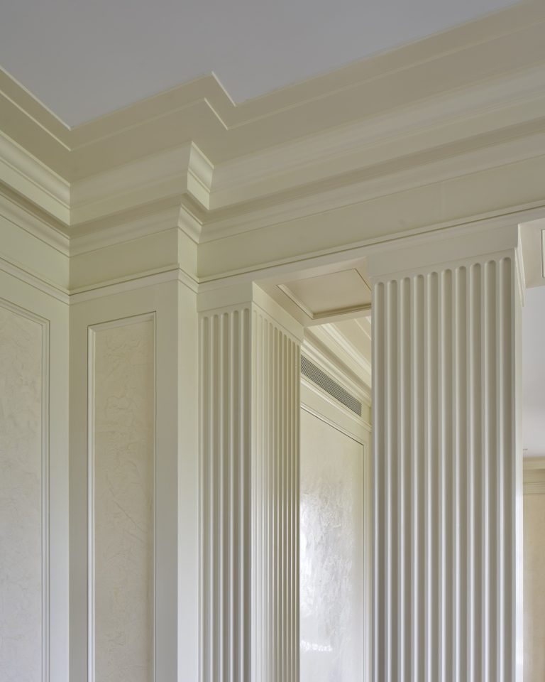 Elegant interior architecture inspired by a Fifth Avenue Renovation in New York, featuring cream-colored walls with intricate molding and trim. Fluted columns frame a doorway as soft, natural light highlights the raised panel details and textured finishes.