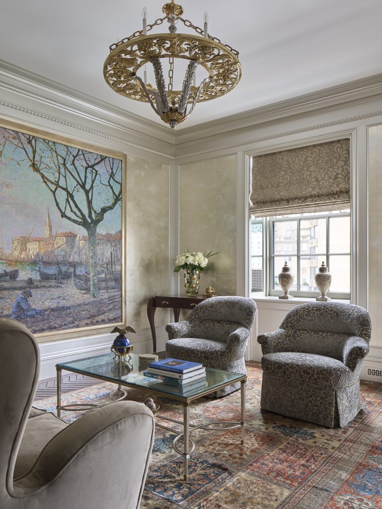 Elegant New York living room with ornate gold chandelier, large impressionist painting, patterned armchairs, glass coffee table with books, a floral arrangement on a console table, classic rug, and window with Roman shade—part of a Fifth Avenue renovation.
