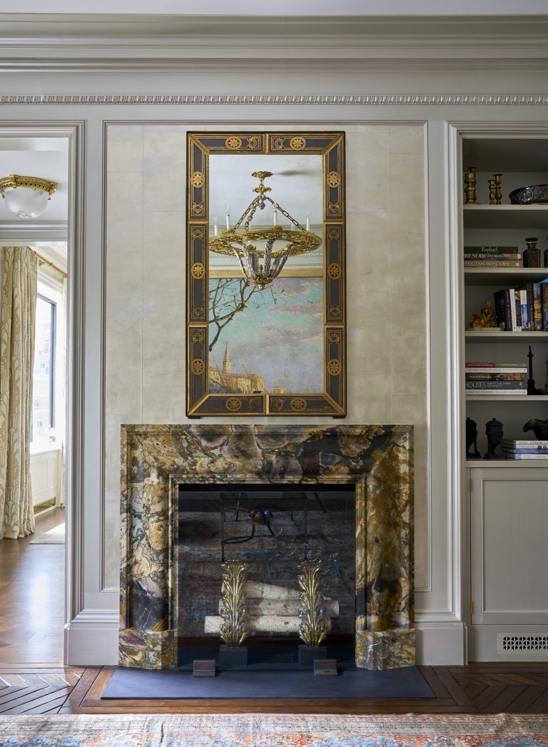 As part of a Fifth Avenue Renovation, a marble fireplace with rich brown and gold veining is set in a cream-paneled wall. Above it hangs a framed mirror reflecting a chandelier, while built-in shelves to the right display books and decorative objects.