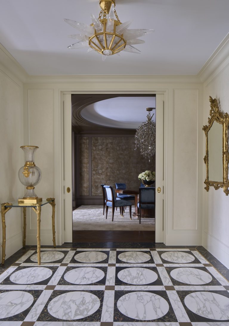 Elegant hallway with a black and white marble floor, golden framed mirror, and ornate glass vase on a golden stand showcases this New York Fifth Avenue Renovation. Beyond the doorway, blue chairs and a crystal chandelier grace the dining room.