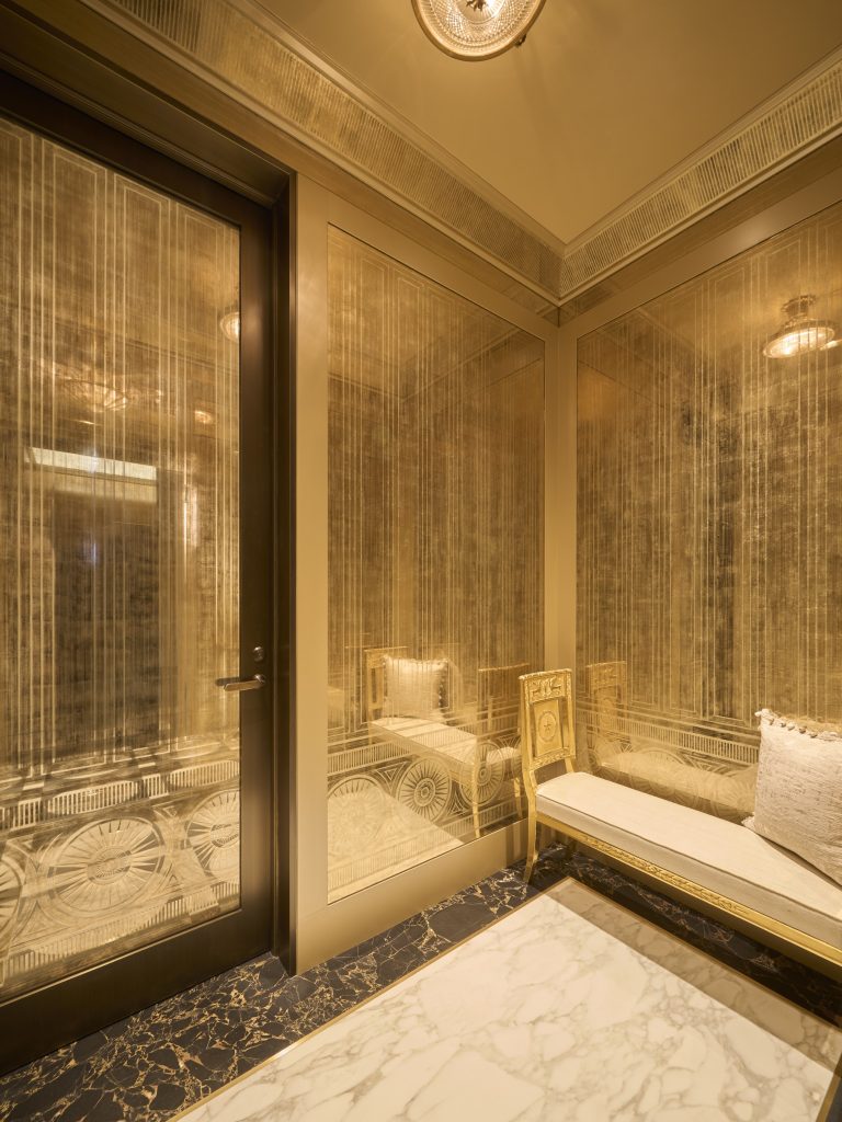 A luxurious, small room inspired by a Fifth Avenue Renovation in New York, with gold-toned glass walls, ornate light fixtures, a marble floor, and a gold-trimmed bench. Warm, elegant lighting creates a lavish atmosphere.