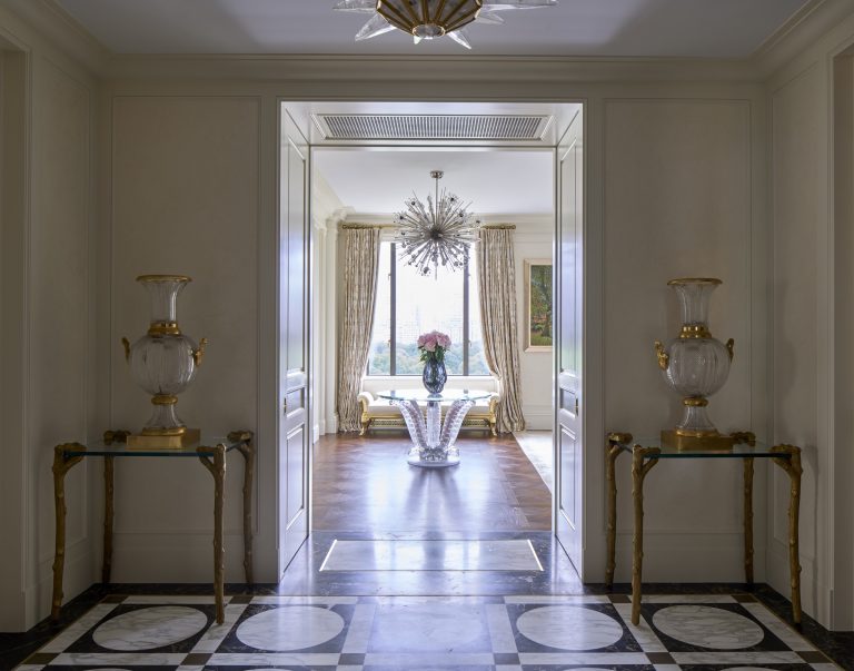 Elegant hallway from a Fifth Avenue Renovation featuring a black-and-white checkered marble floor, ornate vases on glass tables, and a doorway leading to a bright room with chandelier and floral arrangement—a classic touch of New York sophistication.