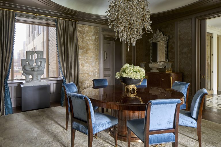 Elegant dining room in a Fifth Avenue Renovation, featuring a round glossy wood table, six blue velvet chairs, and a cascading crystal chandelier. A white floral arrangement sits beneath large windows with blue curtains and an ornate mirror.