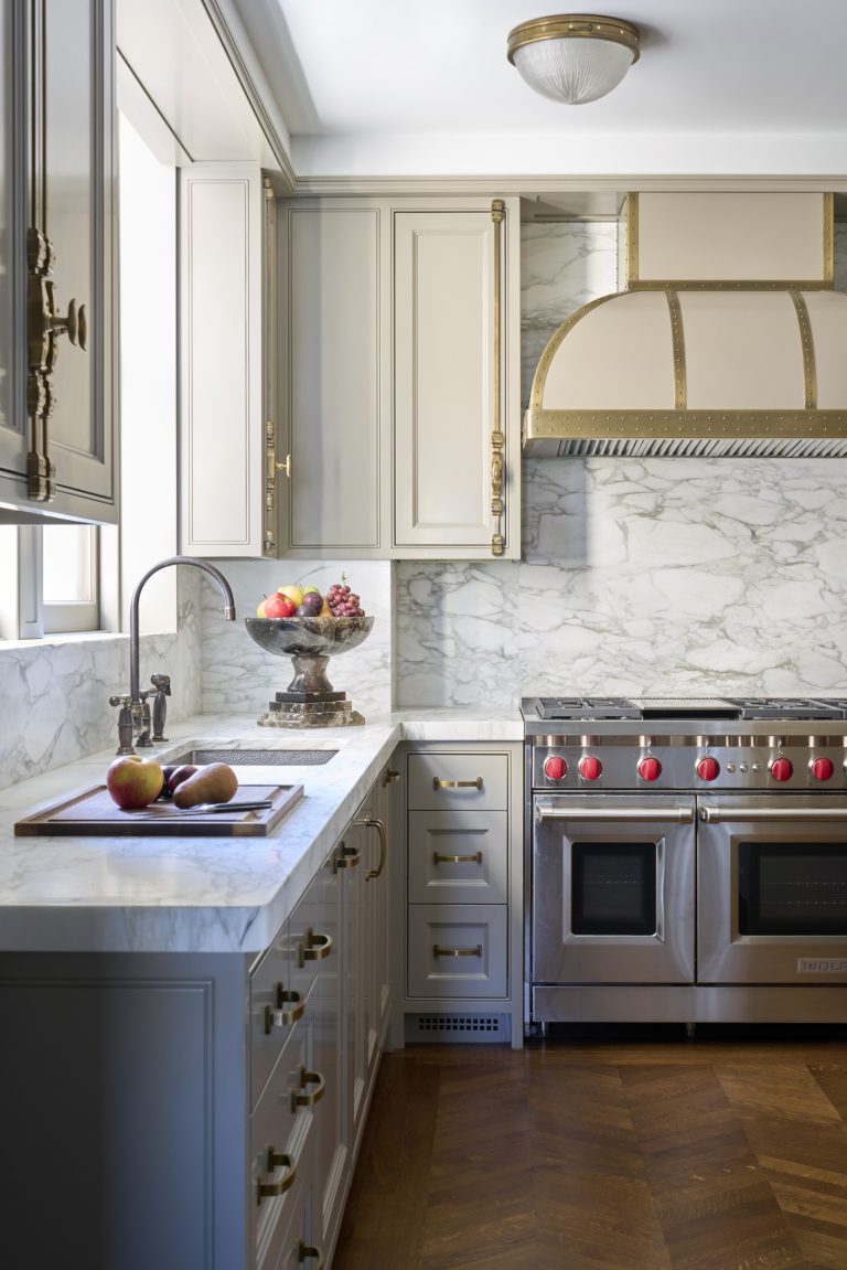 Elegant kitchen with marble countertops and backsplash, stainless steel double oven with red knobs, gold-trimmed range hood, brass hardware, and wood floor—part of a stunning New York Fifth Avenue Renovation. A fruit bowl and apples on a cutting board add charm.
