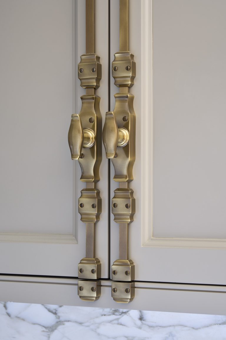 Two beige cabinet doors with ornate, elongated brass handles and decorative rectangular plates evoke Fifth Avenue Renovation style. A white marble countertop with grey veining adds New York elegance to the scene.