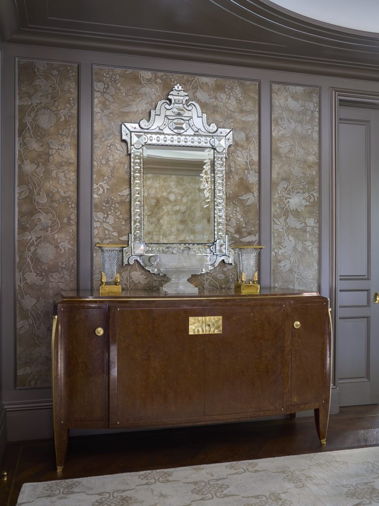 A mirrored cabinet stands against ornate, floral-patterned wallpaper in this Fifth Avenue Renovation. Above it hangs a Venetian-style mirror, while two glass vases with gold bases sit atop, echoing classic New York elegance.