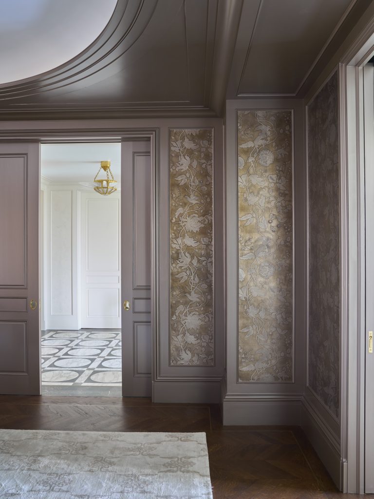 A luxurious hallway in a New York Fifth Avenue Renovation features brown paneled walls with gold floral wallpaper, a pale area rug on dark wood floors, and an open doorway to a bright room with patterned tile and a gold chandelier.