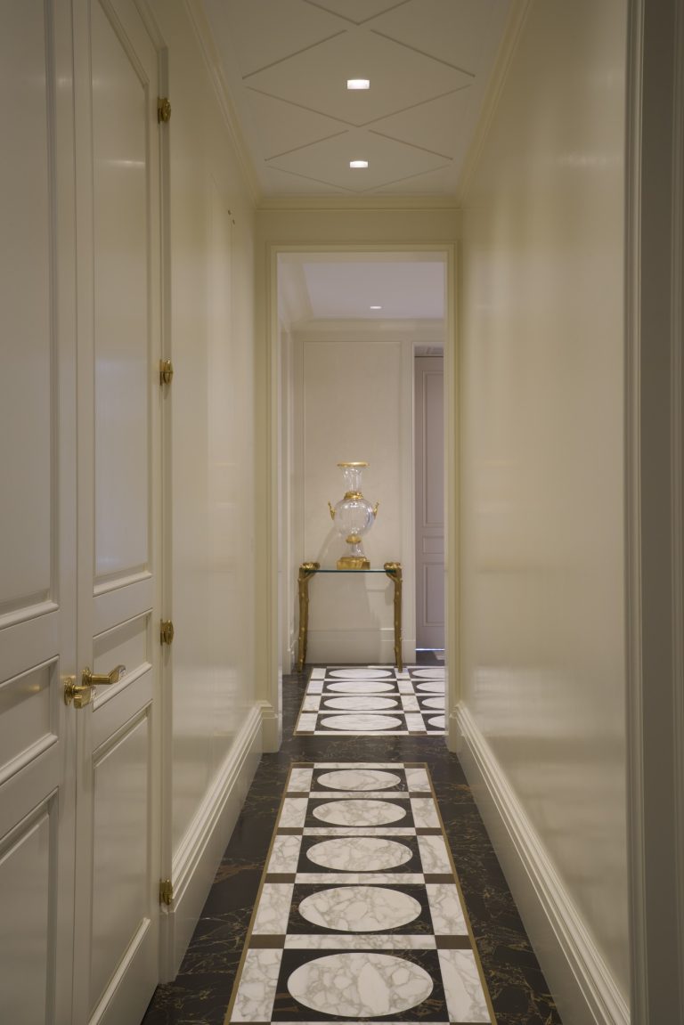 A narrow, elegant hallway inspired by a Fifth Avenue Renovation in New York features cream walls, paneled doors, and a gold-trimmed table with a decorative urn. Black, white, and gray geometric marble patterns line the floor under soft recessed lighting.