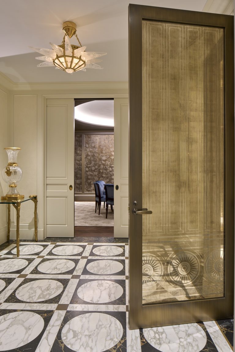 Elegant entryway inspired by a Fifth Avenue Renovation, featuring a large glass and metal door. Marble tile floor has black and white circular patterns. Beige paneled walls, gold-accented side table, and chic dining room with blue chairs beyond.