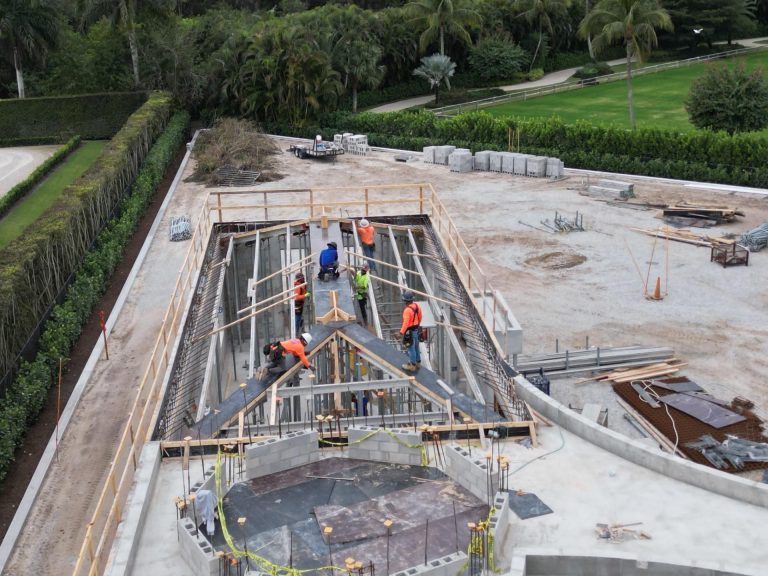 Aerial view of construction workers in orange and blue safety vests building a large concrete structure with steel beams, surrounded by equipment, materials, and greenery. The area is partially paved with trees and grass in the background.