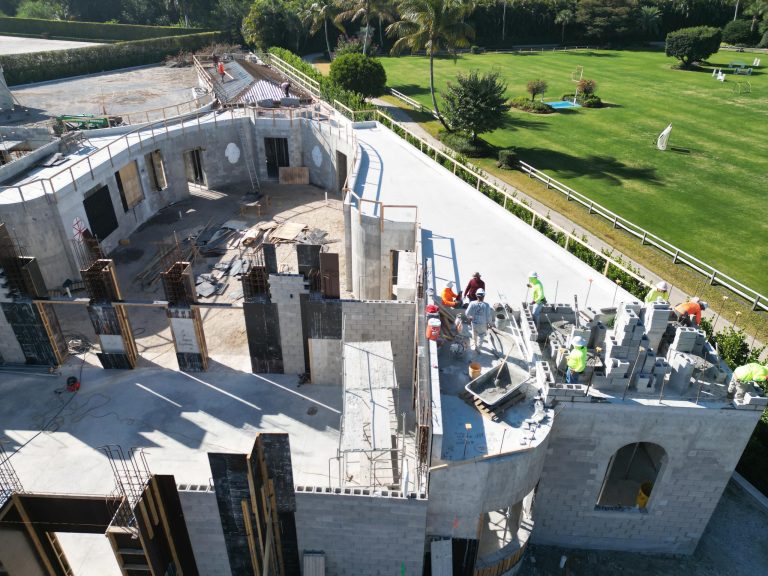A group of construction workers build a large concrete structure with curved walls, scaffolding, and construction materials. The site borders a green lawn with palm trees, a fence, and landscaping visible in the background.