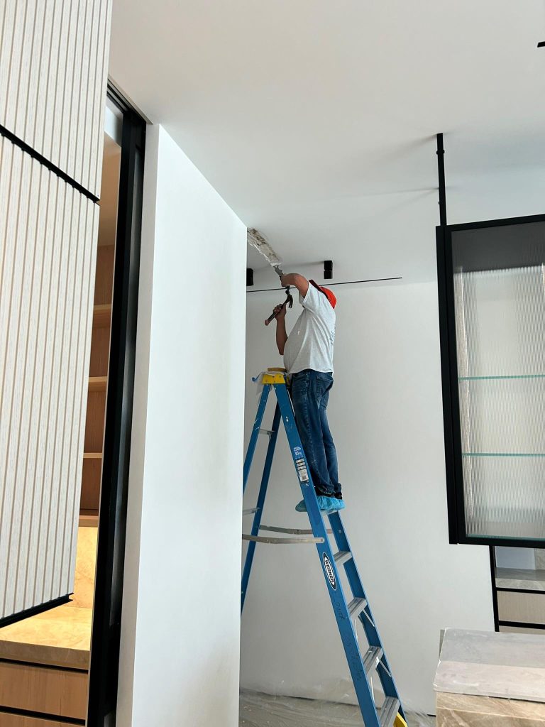 A person stands on a blue ladder, wearing jeans and a white shirt with red sleeves, installing or repairing something on the white ceiling of a modern, minimalistic room with a partially open wall and glass shelving.