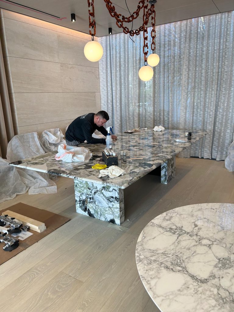A man cleans a large, rectangular marble table with gray veins in a modern room. Large round pendant lights with brown chain links hang above. Sheer curtains cover the window, and unpacked items are scattered on light wooden floors and surrounding chairs.