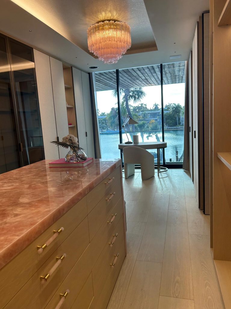 A modern walk-in closet with light wood cabinets, brass handles, and a pink marble island. A glass vase with flowers sits on the island. At the far end, floor-to-ceiling windows reveal a water view, palm trees, and a small round table with two chairs.