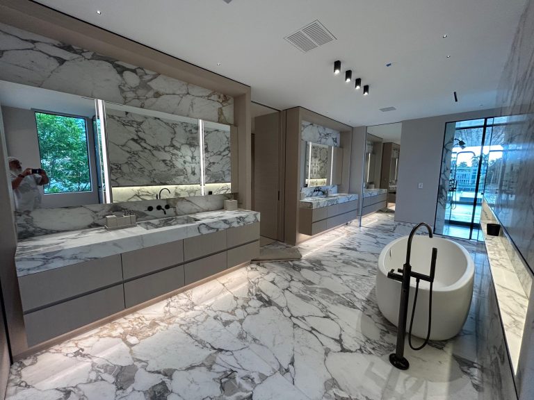 A modern bathroom with marble floors and walls, two large vanities with mirrors, a white freestanding bathtub with a black faucet, and floor-to-ceiling windows letting in natural light. A person is partially visible in the mirror taking a photo.