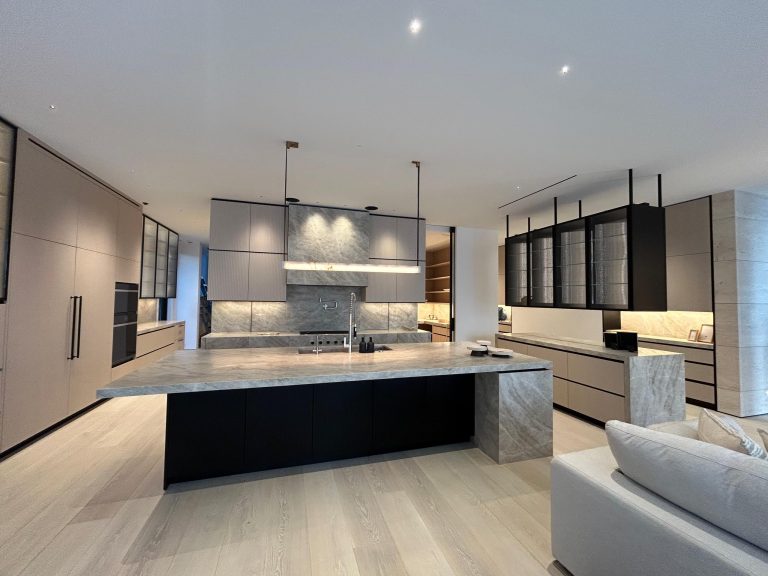 Modern kitchen with a large marble island, black base, and built-in sink. Flat-panel cabinets in beige and black flank the space. Marble backsplash, open shelves, and under-cabinet lighting create a sleek, minimalist look. Light wood flooring and neutral tones throughout.