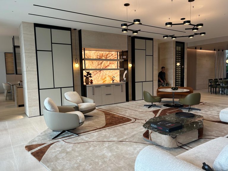 A modern, spacious living room with beige walls, geometric-patterned rugs, marble and wood furniture, plush armchairs, a circular table, and a unique bookshelf. Pendant lights hang from the ceiling. A person stands near a wall in the background.