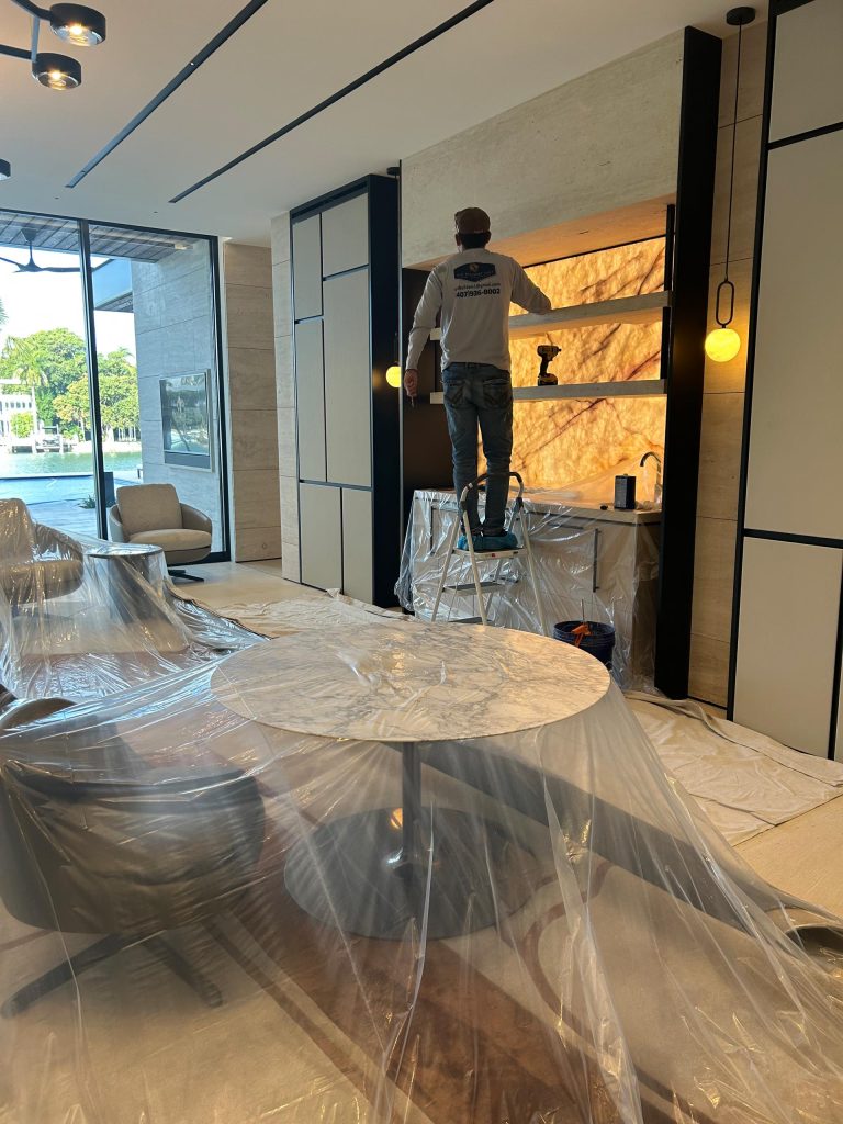 A worker stands on a ladder using a power tool to install or fix shelving in a modern, well-lit room. Furniture and the floor are covered in plastic sheets for protection. Shelves are backlit and the space features contemporary decor with large windows.